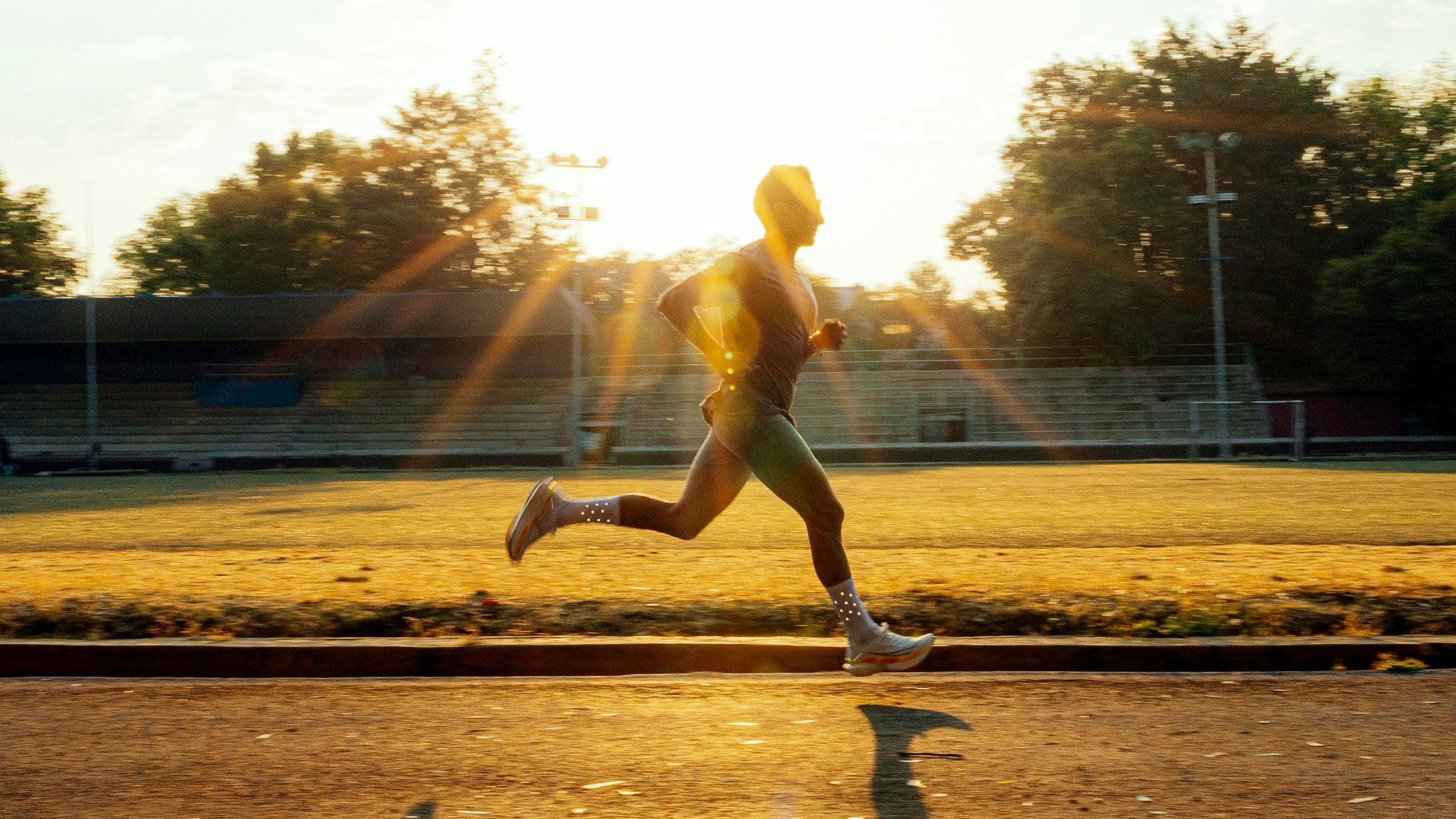 A runner at full stride on a track, lit from behind by a low sun that throws a long shadow across the lane.